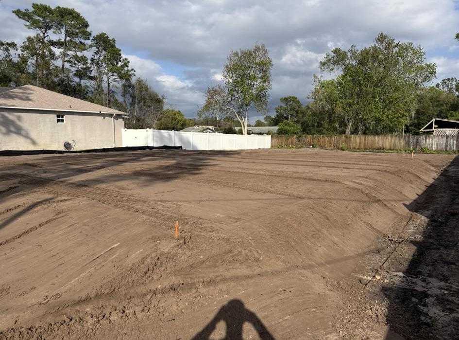 Freshly graded residential lot with smooth dirt surface next to home with white vinyl fence in Deltona