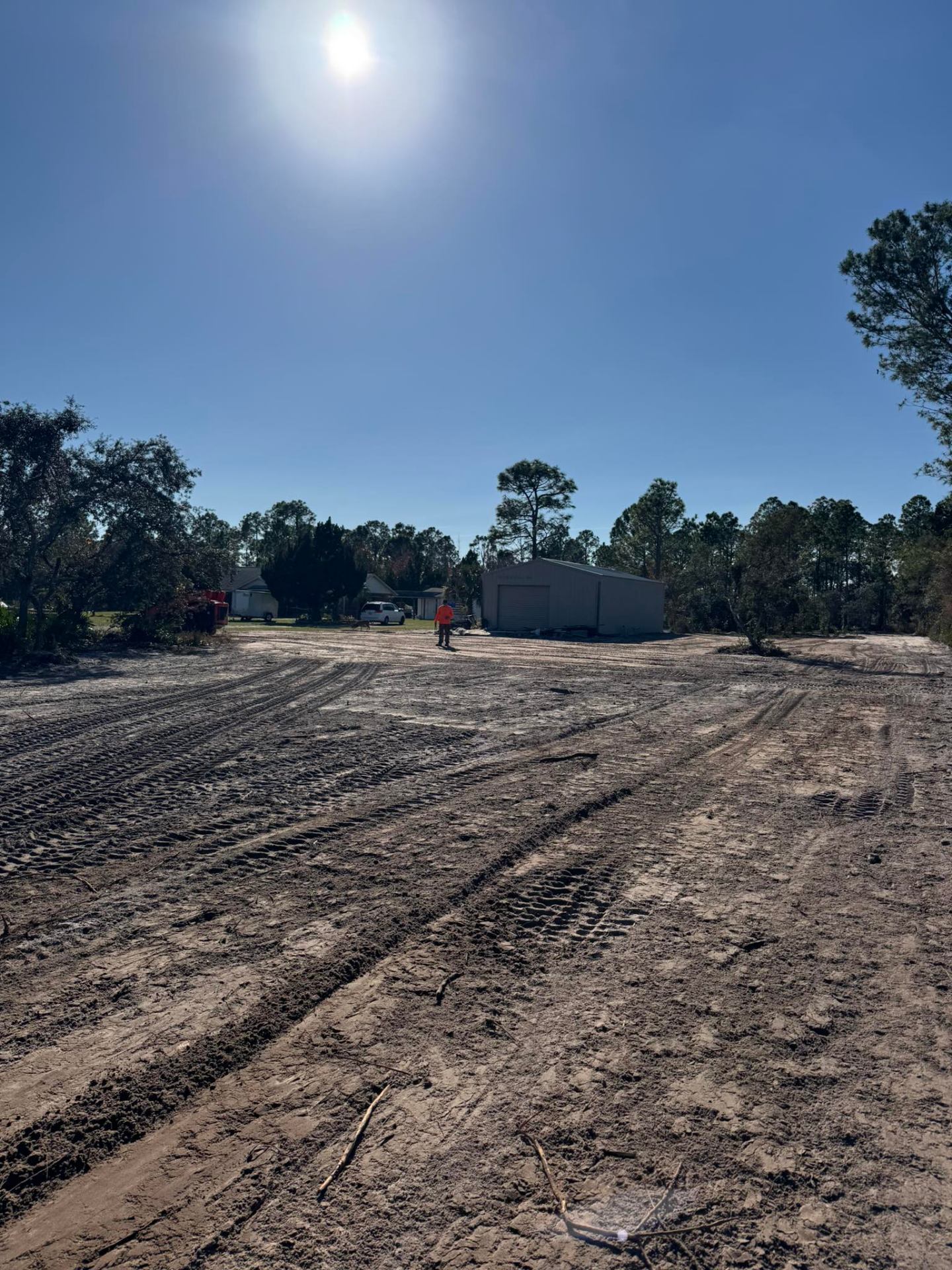Worker in orange shirt walking across freshly graded lot with metal building and bright sun in Deltona