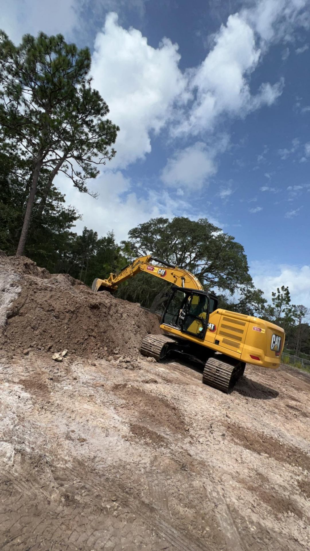 CAT 320 excavator on large dirt pile with oak trees and cloudy sky on Florida excavation site